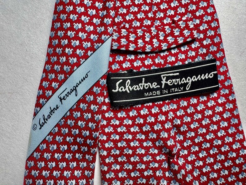 Puede incluir: Una corbata roja con un patr&oacute;n repetitivo de peque&ntilde;os elefantes blancos y azul claro. La corbata incluye una etiqueta de tela azul claro y una etiqueta negra con el texto Salvatore Ferragamo MADE IN ITALY impreso en blanco.