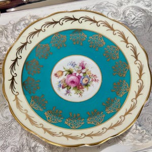 May include: A vintage teacup saucer with a teal center featuring gold filigree and a floral bouquet. The saucer has a cream-colored rim with gold accents and a scalloped edge. The teacup saucer is a classic piece of tableware.
