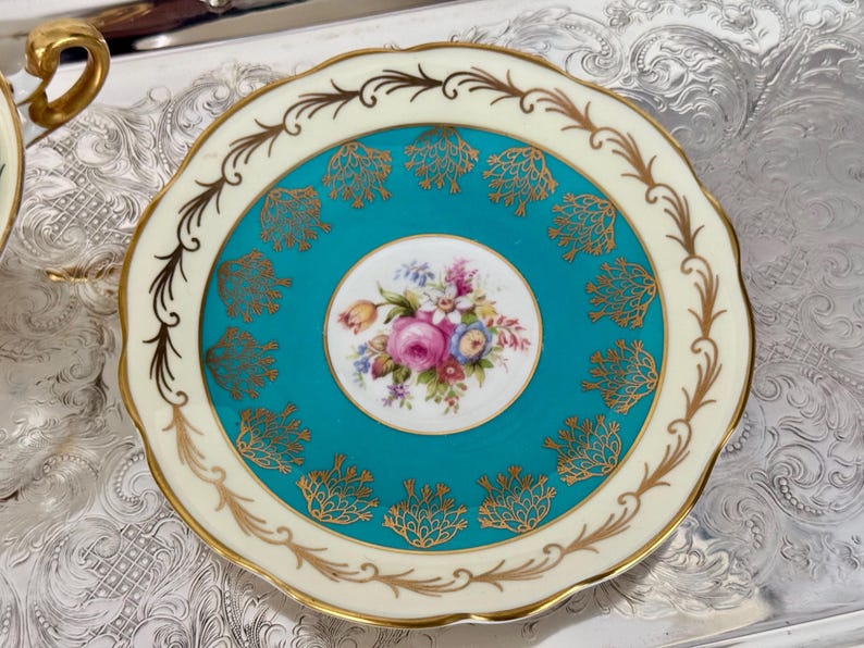 May include: A vintage teacup saucer with a teal center featuring gold filigree and a floral bouquet. The saucer has a cream-colored rim with gold accents and a scalloped edge. The teacup saucer is a classic piece of tableware.