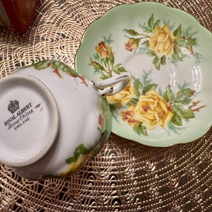 May include: A vintage Royal Albert Bone China teacup and saucer set with a yellow rose floral pattern. The saucer is a pale green color with a scalloped edge. The teacup has a white base with a gold trim.