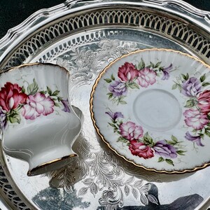 May include: A white teacup and saucer set with a floral design in shades of red, pink, and purple, accented with gold trim. The set is placed on a silver serving tray with an ornate, embossed pattern. A classic tea set.
