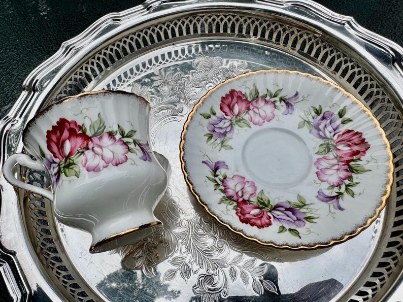 May include: A white teacup and saucer set with a floral design in shades of red, pink, and purple, accented with gold trim. The set is placed on a silver serving tray with an ornate, embossed pattern. A classic tea set.