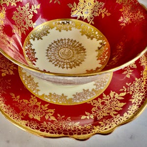 May include: A red and gold teacup and saucer set. The teacup and saucer have a scalloped edge and are decorated with gold floral patterns. The teacup is nested inside the saucer, and the interior of the teacup is a cream color with a gold design.