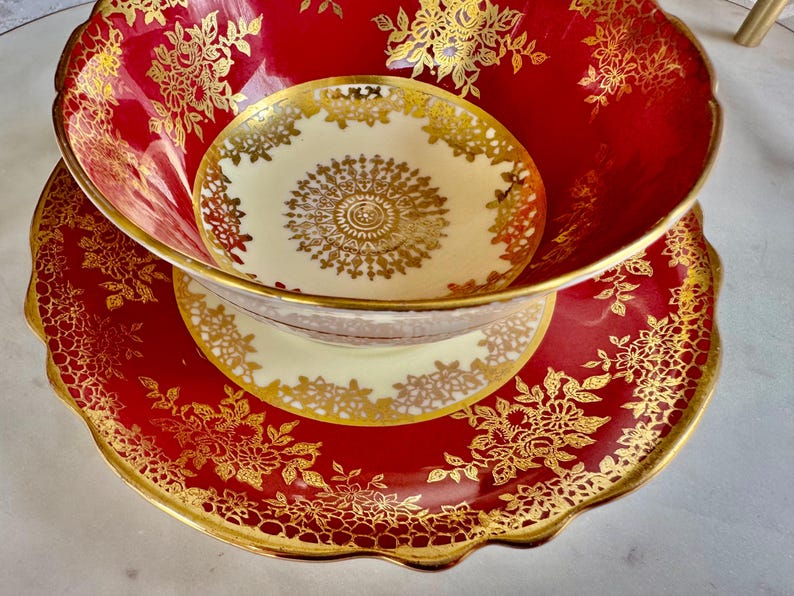 May include: A red and gold teacup and saucer set. The teacup and saucer have a scalloped edge and are decorated with gold floral patterns. The teacup is nested inside the saucer, and the interior of the teacup is a cream color with a gold design.