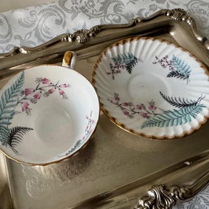 Puede incluir: Un juego de taza y platillo de t&eacute; vintage con borde dorado, decorado con flores rosas y hojas de helecho en verde azulado y negro. El juego descansa sobre una bandeja plateada, evocando una sensaci&oacute;n cl&aacute;sica y elegante.
