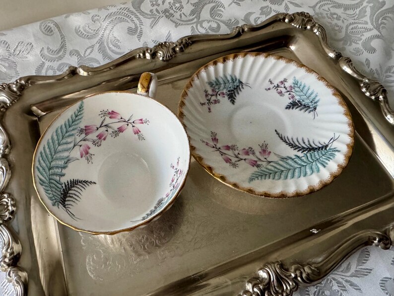 Puede incluir: Un juego de taza y platillo de t&eacute; vintage con borde dorado, decorado con flores rosas y hojas de helecho en verde azulado y negro. El juego descansa sobre una bandeja plateada, evocando una sensaci&oacute;n cl&aacute;sica y elegante.