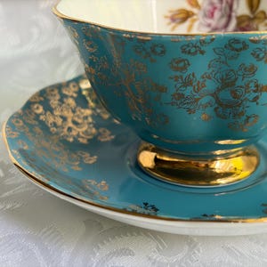 Może przedstawiać: Zestaw filiżanki do herbaty ze spodkiem z turkusowej porcelany z metalicznym złotym wzorem kwiatowym. Filiżanka posiada złocony brzeg oraz złotą podstawę, a jej wnętrze zdobi niewielki motyw kwiatowy na białym tle.