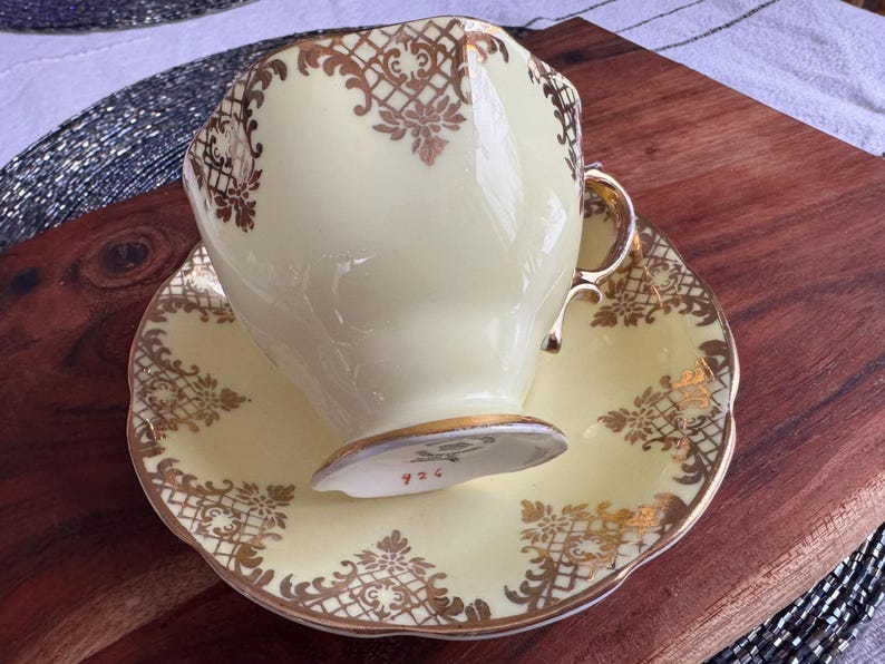 May include: A yellow teacup and saucer with a gold floral pattern. The cup has a gold rim and the saucer has a scalloped edge.