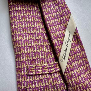 Puede incluir: Una corbata morada con un patr&oacute;n repetitivo de peque&ntilde;as figuras de animales de color claro. La corbata incluye una etiqueta de tela color crema cosida en la parte posterior que muestra el texto Salvatore Ferragamo en letra cursiva negra.
