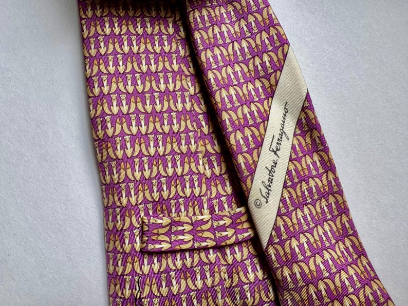 Puede incluir: Una corbata morada con un patr&oacute;n repetitivo de peque&ntilde;as figuras de animales de color claro. La corbata incluye una etiqueta de tela color crema cosida en la parte posterior que muestra el texto Salvatore Ferragamo en letra cursiva negra.
