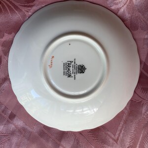 May include: A white bone china saucer with a scalloped edge. The underside features a black stamp reading BY APPOINTMENT TO HER MAJESTY THE QUEEN CHINA POTTERS PARAGON FINE BONE CHINA ENGLAND REGD and a handwritten orange number A4339.