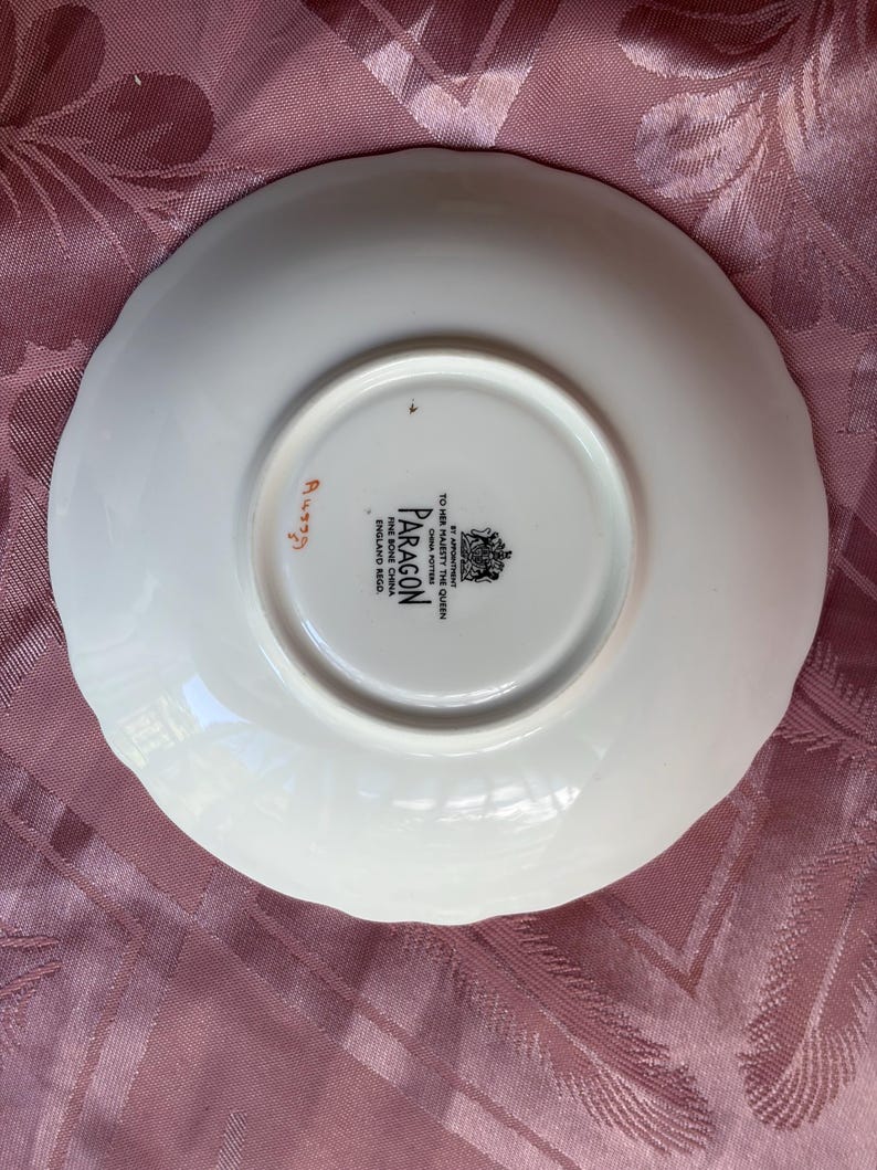 May include: A white bone china saucer with a scalloped edge. The underside features a black stamp reading BY APPOINTMENT TO HER MAJESTY THE QUEEN CHINA POTTERS PARAGON FINE BONE CHINA ENGLAND REGD and a handwritten orange number A4339.