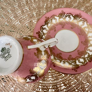 May include: A pink teacup and saucer set with gold filigree detailing. The teacup has a white interior and a white handle. The saucer is round with a white center. The set is made of fine bone china.