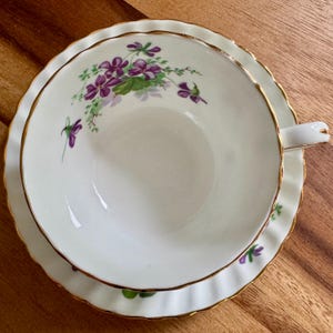 K&ouml;nnte beinhalten: Wei&szlig;es Tee-Set mit gewelltem Rand und goldenem Rand. Die Tasse und Untertasse haben ein Blumendesign mit lila Blumen und gr&uuml;nen Bl&auml;ttern. Das Set steht auf einer Holzoberfl&auml;che.