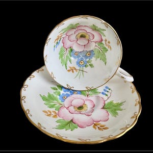 May include: A white teacup and saucer set with a floral design. The cup and saucer are decorated with pink and blue flowers, green leaves, and gold trim.