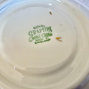 Może przedstawiać: Biały spodek Royal Grafton Bone China z lekko podniesioną krawędzią. Na środku zielony napis "Royal Grafton Bone China Made in England". Spodek ma błyszczące wykończenie.