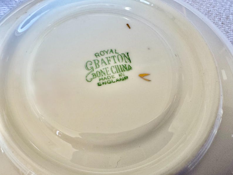 Może przedstawiać: Biały spodek Royal Grafton Bone China z lekko podniesioną krawędzią. Na środku zielony napis "Royal Grafton Bone China Made in England". Spodek ma błyszczące wykończenie.