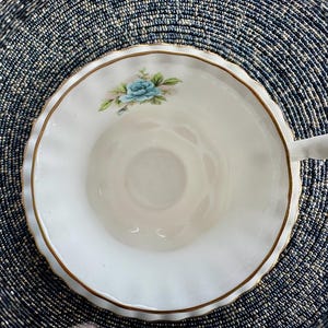 May include: A white teacup with a gold rim and a blue floral design. The teacup has a scalloped edge and a handle.