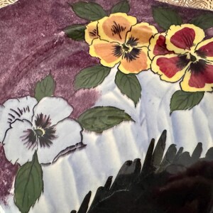 May include: A decorative ceramic plate with a floral design. The plate features hand-painted pansies in shades of yellow, red, and white, with green leaves. The background is a mix of purple and white, with a black accent.