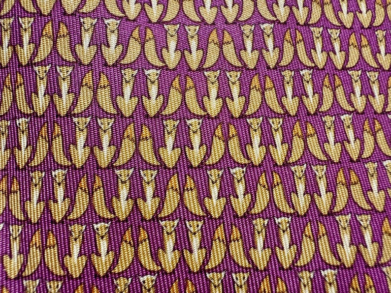 Puede incluir: Una tela de color p&uacute;rpura con un patr&oacute;n repetitivo de peque&ntilde;os zorros estilizados de color marr&oacute;n dorado. Los zorros est&aacute;n representados en posici&oacute;n sentada, dispuestos en filas uniformes sobre el material texturizado.