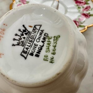 Puede incluir: Primer plano de la base de una taza de t&eacute; Salisbury Bone China blanca, con el logotipo de la marca y "ENGLAND" impreso. La taza tiene una forma curvada. Un platillo floral est&aacute; en el fondo.