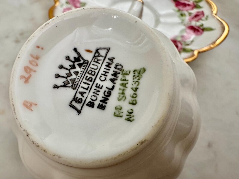 Puede incluir: Primer plano de la base de una taza de t&eacute; Salisbury Bone China blanca, con el logotipo de la marca y "ENGLAND" impreso. La taza tiene una forma curvada. Un platillo floral est&aacute; en el fondo.