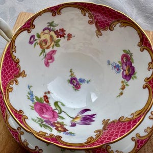 May include: A white teacup with a pink and gold rim, featuring a floral design. The cup has a scalloped edge and a handle. The interior of the cup is white, and the floral design includes pink, purple, and yellow flowers.
