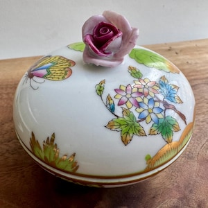 May include: A white porcelain lidded dish with a floral and butterfly design. The lid features a pink rose with a burgundy center.