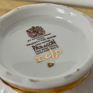 May include: Close-up of the bottom of a white Paragon fine bone china teacup. The cup's interior features a gold rim and a printed emblem with text that reads "BY APPOINTMENT TO HER MAJESTY THE QUEEN CHINA POTTERS PARAGON FINE BONE CHINA ENGLAND REGD".