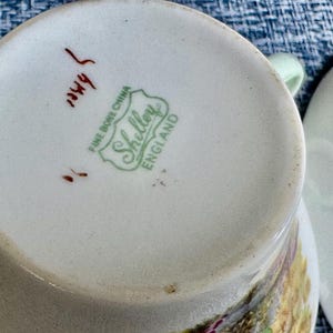 May include: The underside of a white fine bone china teacup featuring a green stamp that reads Shelley Fine Bone China England. There is also a handwritten red mark on the base. A portion of a matching white saucer is visible to the right side.