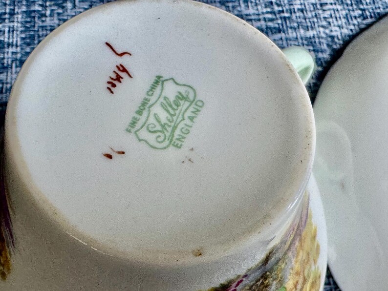 May include: The underside of a white fine bone china teacup featuring a green stamp that reads Shelley Fine Bone China England. There is also a handwritten red mark on the base. A portion of a matching white saucer is visible to the right side.