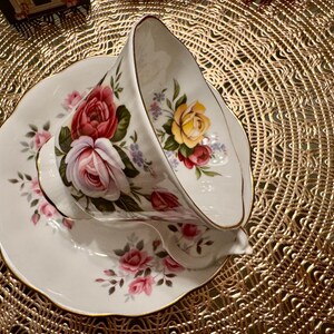 May include: A white teacup and saucer with a floral pattern featuring pink, red, and yellow roses. The cup is tilted on its side, and the saucer has a gold rim.