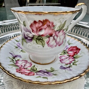 May include: A white teacup and saucer set with a floral design in shades of pink, purple, and green. The edges of the cup and saucer are trimmed with gold. The teacup has a curved handle.