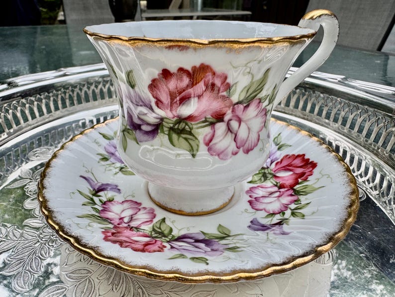 May include: A white teacup and saucer set with a floral design in shades of pink, purple, and green. The edges of the cup and saucer are trimmed with gold. The teacup has a curved handle.