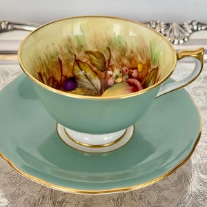 May include: A vintage teacup and saucer set. The cup and saucer are a soft teal color with gold trim. The inside of the cup features a hand-painted design of fruit and foliage in shades of red, yellow, and purple. The set is displayed on a silver tray.