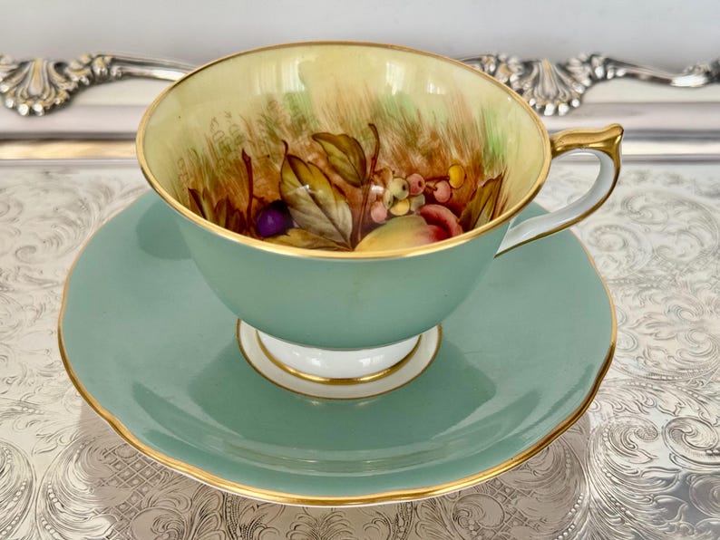 May include: A vintage teacup and saucer set. The cup and saucer are a soft teal color with gold trim. The inside of the cup features a hand-painted design of fruit and foliage in shades of red, yellow, and purple. The set is displayed on a silver tray.