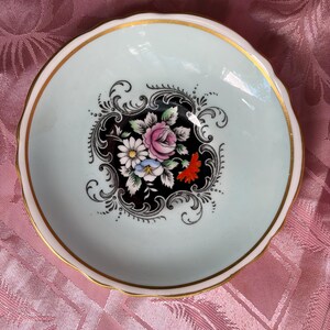 May include: A small, round porcelain dish with a pale mint green rim and a gold-trimmed scalloped edge. The center features a black decorative medallion containing a colorful floral arrangement of pink, white, blue, and orange flowers with green leaves.