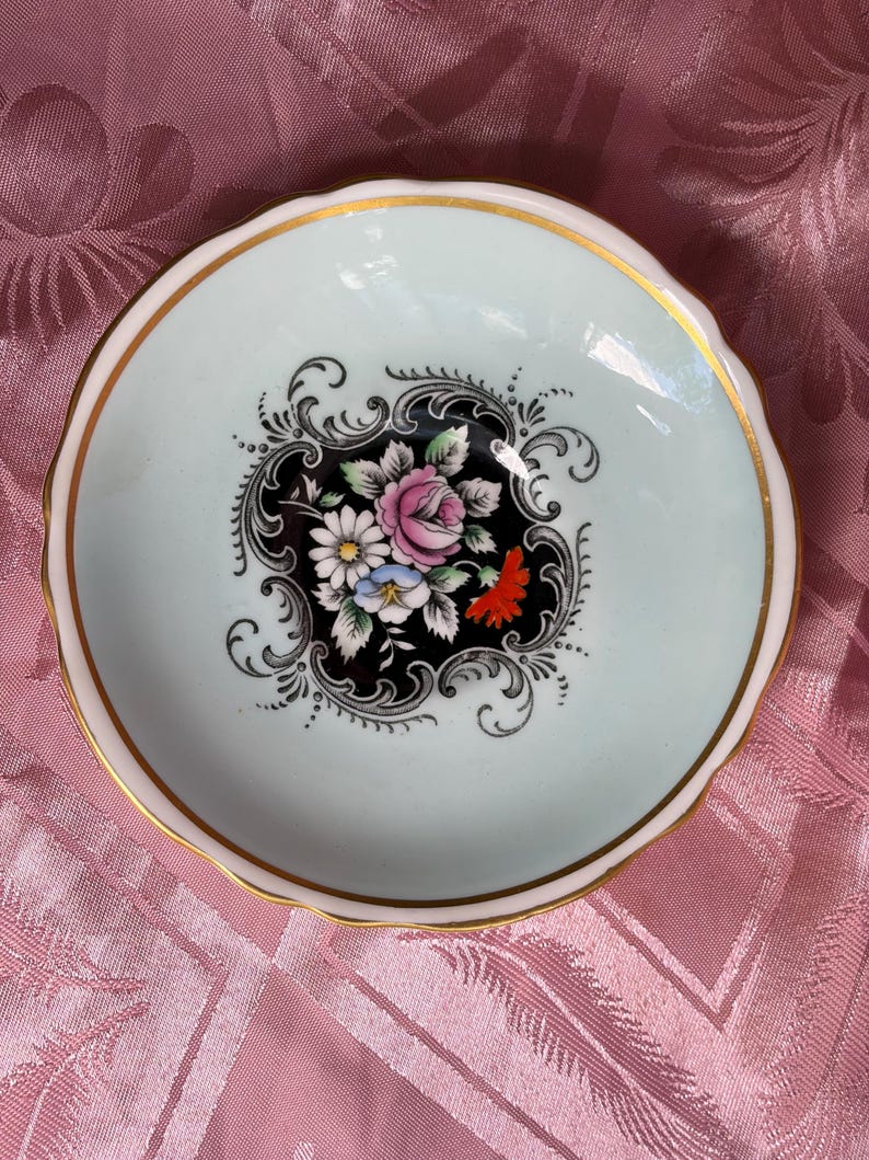 May include: A small, round porcelain dish with a pale mint green rim and a gold-trimmed scalloped edge. The center features a black decorative medallion containing a colorful floral arrangement of pink, white, blue, and orange flowers with green leaves.