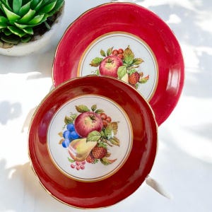 May include: A red teacup and saucer set with a gold rim and a white center featuring a colorful fruit design. The fruit design includes apples, pears, grapes, and strawberries.
