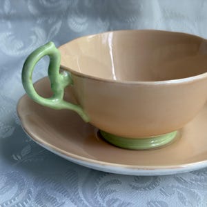May include: A peach-colored porcelain teacup and saucer set featuring a light green handle and a matching light green base. The teacup has a smooth, rounded bowl shape with a scalloped rim, while the saucer has a matching scalloped edge.