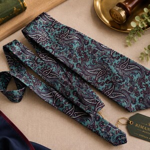 K&ouml;nnte beinhalten: Eine blaugr&uuml;ne Krawatte mit einem detaillierten Paisley-Muster in dunklen Violett-, Schwarz- und Wei&szlig;t&ouml;nen. Die Krawatte wird von einem dunkelgr&uuml;nen rechteckigen Etikett mit goldenem Text begleitet, der Rimantique Boutique Vintage Finds lautet.