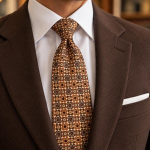 May include: A brown textured blazer with a white dress shirt and a brown tie featuring a repeating geometric floral pattern in shades of tan, black, and dark red. A white pocket square is tucked into the blazer's chest pocket.