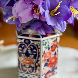 Peut inclure: Un vase hexagonal en c&eacute;ramique avec un motif floral, contenant un bouquet de fleurs violettes. Le vase pr&eacute;sente un motif bleu et blanc avec des accents floraux rouges. Les fleurs ont des &eacute;tamines jaunes.