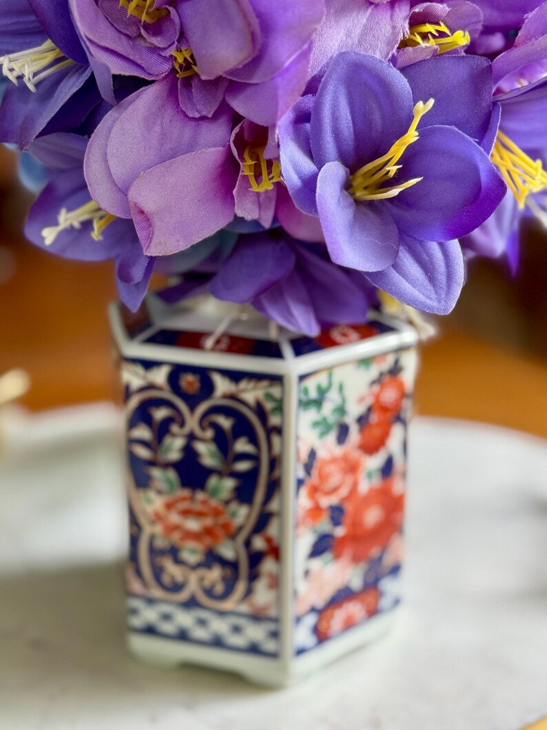 Peut inclure: Un vase hexagonal en c&eacute;ramique avec un motif floral, contenant un bouquet de fleurs violettes. Le vase pr&eacute;sente un motif bleu et blanc avec des accents floraux rouges. Les fleurs ont des &eacute;tamines jaunes.