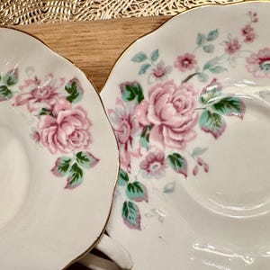 May include: Two white porcelain teacups with a floral pattern. The cups have a gold trim and feature pink roses and green leaves.