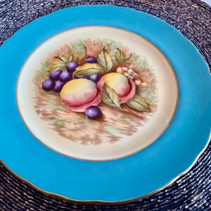 May include: A round, turquoise-colored plate with a gold rim. The center features a painted still life of peaches and grapes. The fruit is surrounded by green leaves and brown stems, set against a cream-colored background.