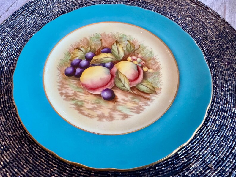May include: A round, turquoise-colored plate with a gold rim. The center features a painted still life of peaches and grapes. The fruit is surrounded by green leaves and brown stems, set against a cream-colored background.