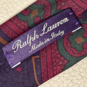 Pode incluir: Uma gravata com um padr&atilde;o paisley em tons de roxo escuro, verde e vermelho. Uma etiqueta de tecido roxa est&aacute; costurada na gravata, exibindo o texto bordado em branco Ralph Lauren e Made in Italy em fonte cursiva.