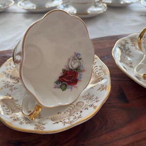 May include: A white teacup and saucer with a gold rim and floral design. The cup features a red rose and white roses with green leaves and blue accents. The saucer has a delicate floral pattern with gold accents.