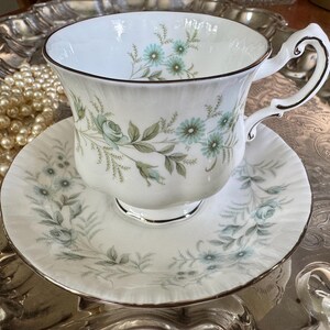 May include: White teacup and saucer set with a floral pattern in shades of blue and green. The teacup has a curved handle and a gold trim. The saucer mirrors the floral design. The set is placed on a decorative silver tray.